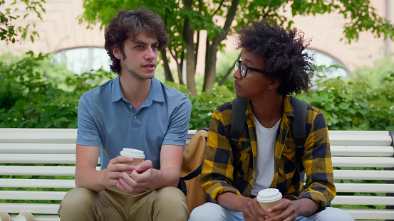 Multiethnic college students sitting on bench relaxing after classes drinking coffee. Afro-american and caucasian male friends talk sitting on park bench with takeaway coffee