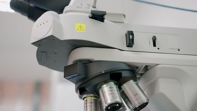 An optical microscope is being operated by the hand of a researcher who is wearing protective gloves. At the same time, the researcher is concurrently placing a polarizer slide into the instrument and adjusting the focal lens to see into minuscule parts.
