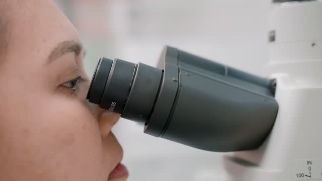 A female scientist adjusts the focus of her eyes on an optical microscope so that she can examine minute details.