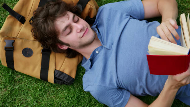 istock Top view of young man lying on grass reading book with head on backpack 1500874882