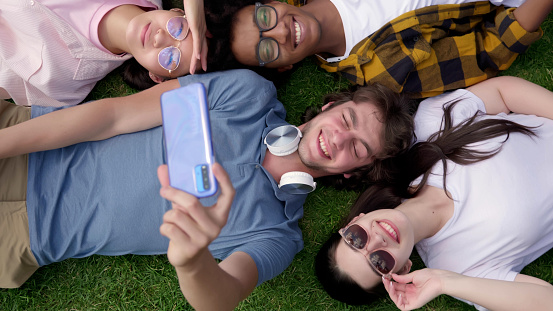 Top view portrait of smiling cheerful multiethnic friends lying together on lawn taking selfie on smartphone. Happy young people lying on grass and taking photo on mobile phone.