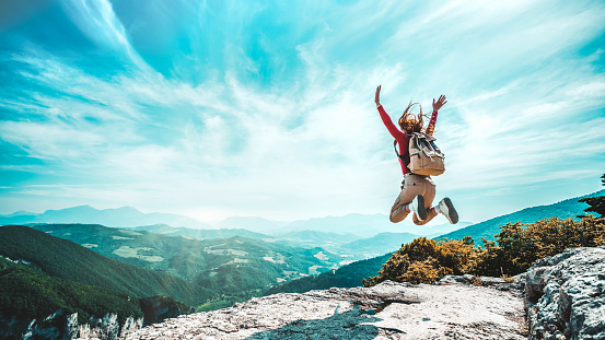 Happy woman with open arms jumping on the top of mountain - Hiker girl with backpack celebrating success outdoor in sunny day - People, success, nature and sport concept
