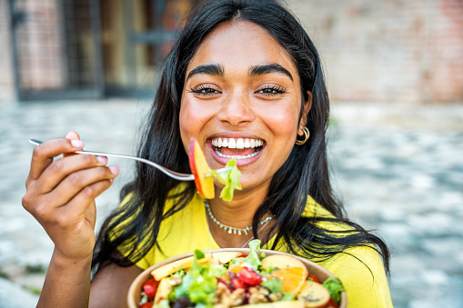 Portrait of young happy african woman eating healthy salad sitting on city street with green fresh ingredients - Asian black student girl having vegan vegetarian lunch break outdoors
