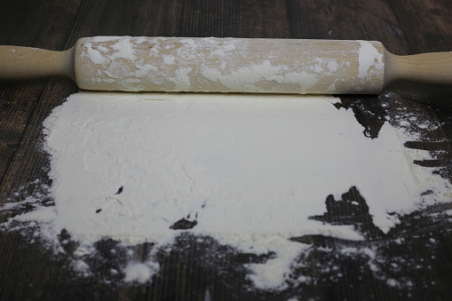 Wooden light rolling pin for dough lies in flour on a dark wooden table. Flour for dough on countertop in kitchen