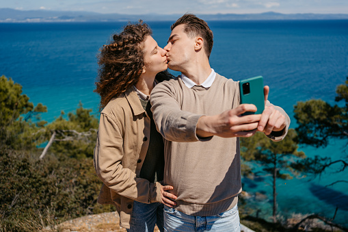 Beautiful young couple kissing and taking selfies on the coast in Greece.