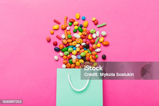 istock shopping paper gift bag in corner full of assorted traditional candies falling out on colored background with copy space. Happy Holidays sale concept 1500897253