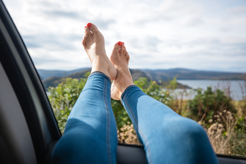 A point of view of an unrecognisable woman with her feet up out of the window of the car. The view is a beauty spot in the Scottish highlands in Loch Torridon