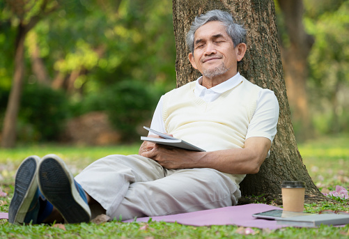 an old man sitting against a tree his eyes closed and relax while reading a book in the park,concept for elderly people activity,hobbies,relaxing,resting,wellbeing