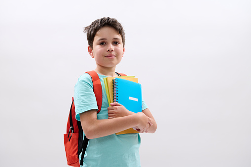 Happy positive teenager smiling looking at camera. Smart schoolboy carrying orange backpack, colorful textbooks and school supplies, isolated on white studio background with copy advertising space
