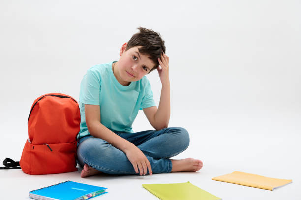 Puzzled doubtful pensive teenager school boy, scratches his head, sitting next to arranged textbooks and orange backpack stock photo
