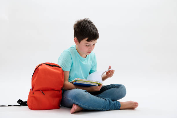 Smart teenager schoolboy, 11-13 years old leafing through his copybook, doing homework, sitting barefoot in lotus pose near his backpack, isolated white studio background. Education. Back to school