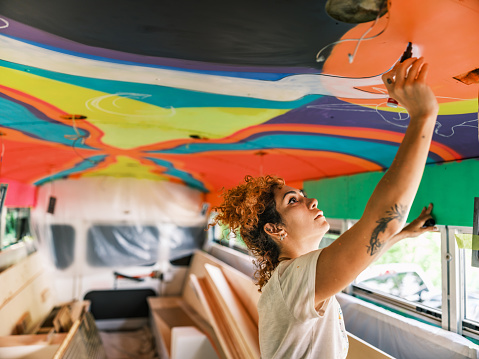 Mid adult Iranian woman artist painting  ceiling mural inside party bus, being converted from traditional school bus.  She is dressed in casual work clothes. Interior of old school bus.