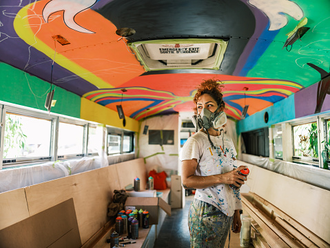 Mid adult Iranian woman artist painting  ceiling mural inside party bus, being converted from traditional school bus.  She is dressed in casual work clothes. Interior of old school bus.