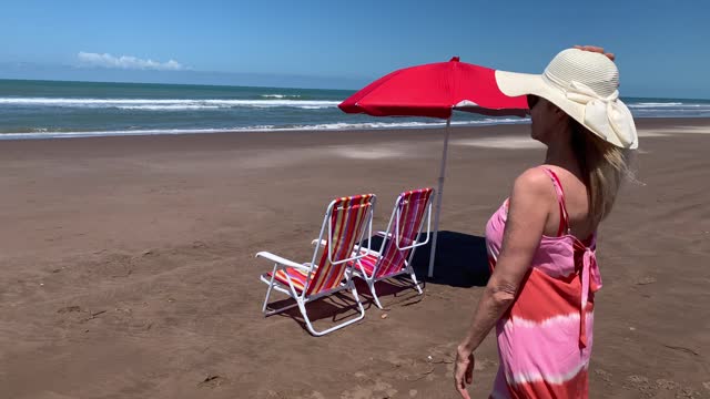 Mature woman enjoying the view while relaxing sitting on a beach chair under an umbrella. Summer concept, video 4k