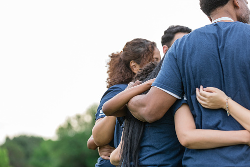 An unrecognizable group of people in matching t-shirts huddle together with their arms around each other.