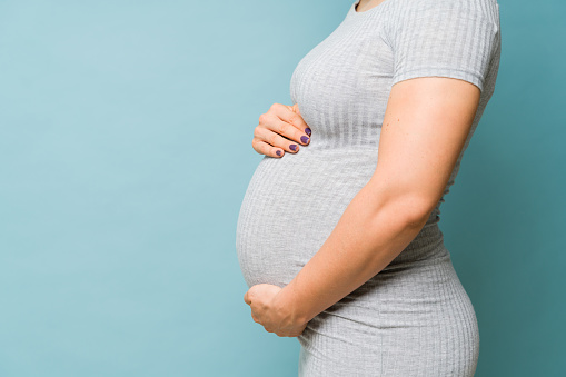 Profile view of the body of a pregnant woman embracing her belly in a studio with a blue background