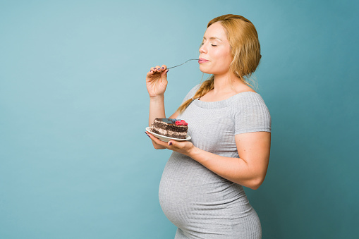 Beautiful pregnant woman giving into her cravings and enjoying a big piece of cake in a studio
