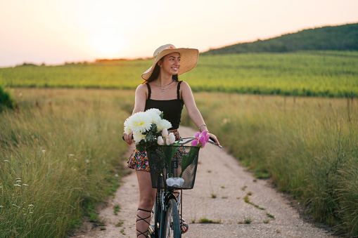 Beautiful girl with a hat enjoying summer day riding a bike. She has flowers in her basket.