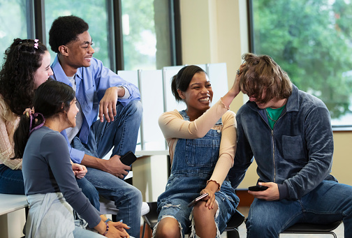 A multiracial group of five high school students having fun together in the school library. The friends are generation Z teenagers, 15 to 18 years old. They are all laughing and smiling, as the African-American girl messes up the long hair of one of the boys.