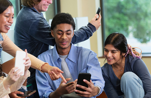 A multiracial group of high school students having fun together in the school library with their smart phones. The friends are generation Z teenagers, 16 to 18 years old. The young African-American man in the middle is showing his mobile phone to two of the girls.