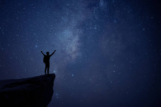Silhouette of young man standing alone on top of mountain and raise both arms enjoying nature on beautiful night sky, star, milky way background. Demonstrates hope and freedom. stock photo