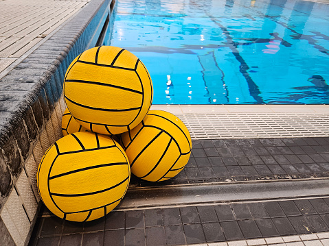 Sports and recreation image of four yellow water polo balls stacked along the edge of an indoor public swimming pool.