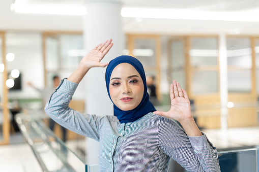 A portrait of a confident Asian professional Muslim female dressed in office attire standing  in office corridor and pose happily in a modern office during coffee break.