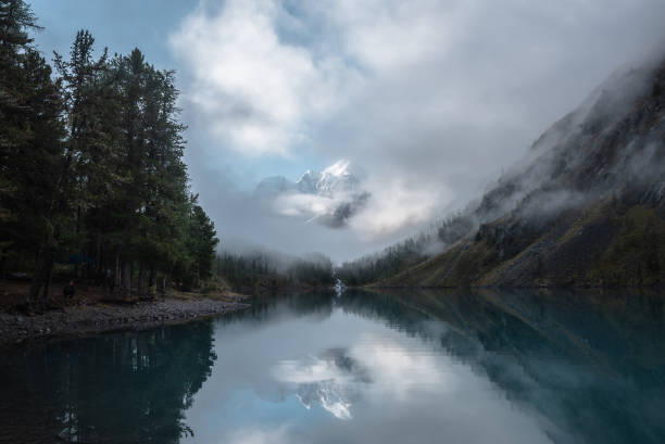 istock Tranquil scenery with snow castle in clouds. Mountain creek flows from forest hills into glacial lake. Snowy mountains in fog clearance, small river and coniferous trees reflected in calm alpine lake. 1501022840