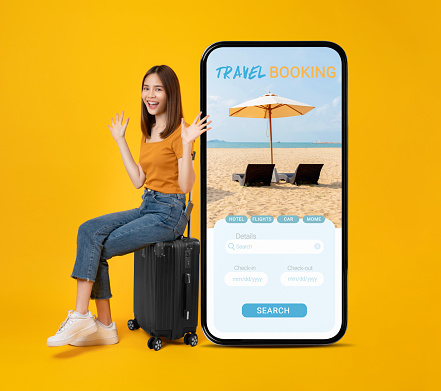 Cheerful young Asian woman sitting on the suitcase and smartphone with application booking flight travel search ticket holiday and hotel on website on yellow background. Concept tourist holiday trip.