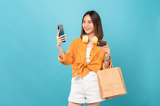 Beautiful Asian woman holding brown blank craft paper shopping bags and showing credit card on blue background.