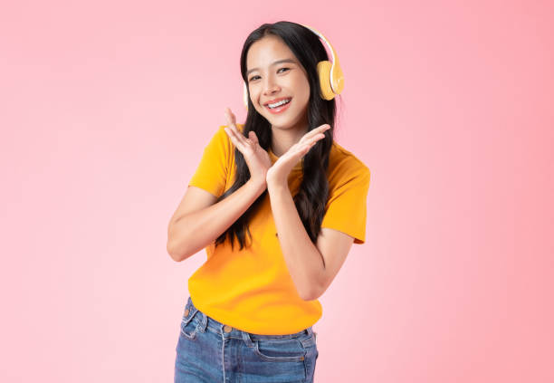 Cheerful young Asian woman in headphones listening to music and enjoy favourite playlist application with dancing on pink background. stock photo