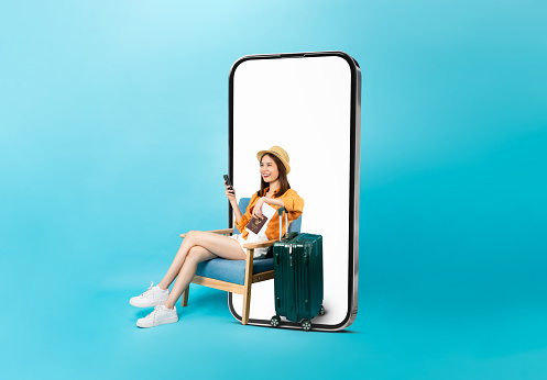Happy Asian woman traveler sitting on the chair and big smartphone with mockup of blank screen, hand holding passport and plane tickets with suitcase isolated on blue background.