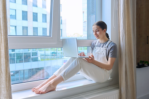 Teenage girl sitting on a windowsill using a laptop. Large panoramic window urban background, young female student using laptop for study leisure communication