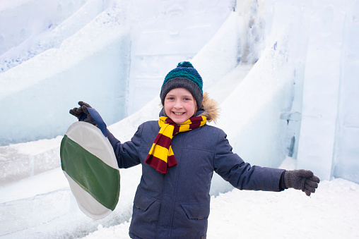 a happy boy holds a  ice skating rink, a joyful child stands in front of an icy city, a castle, a slide, is going to slide down a hill.