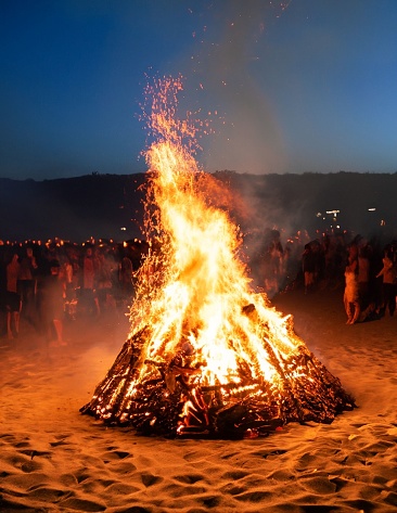 A large beach bonfire is burning brightly on a sandy shoreline, with a group of people gathered around