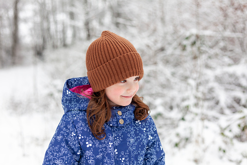 Portrait of a cute little girl in the winter forest. The girl is wearing a warm hat and blue jacket.
