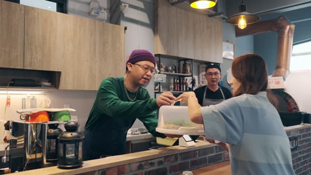 In the restaurant, a woman uses her smartphone to make a payment with the cashier. She scans the QR code using a card reader to complete a convenient contactless payment. At the same time, the cashier hands over the purchased pizza to the customer.