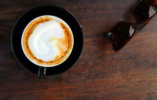 Top view or flat lay of hot cappuccino in black cup with brown sunglasses and on brown wooden background or table with copy space. Refreshment drinking with coffee at cafe shop in the morning time.