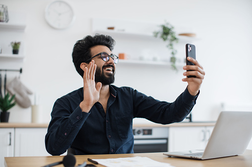 Smiling bearded adult in glasses showing hello gesture while lifting smartphone on eye level at office desk in kitchen. Joyous indian businessman greeting video call participants during working time.