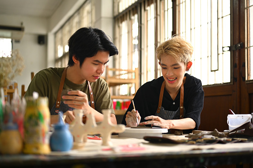 Cheerful young asian gay couple painting ceramics, creating handmade earthenware in workshop.