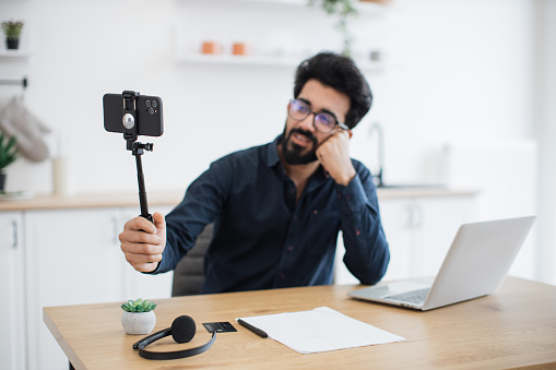 Focus on modern cell phone placed in tripod being held by tired indian man in glasses sitting at desk with laptop. Professional consultant live streaming detailed report from distant workplace.