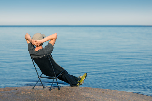 A man is sitting in a camping chair by the ocean looking at the horizon. The background is blurred.