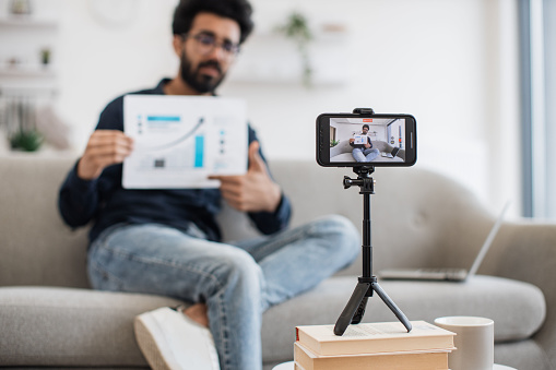 Handsome indian guy in casual clothes sitting on couch with graph financial report in hands while recording video on modern smartphone. Successful financier talking about statistics on camera.