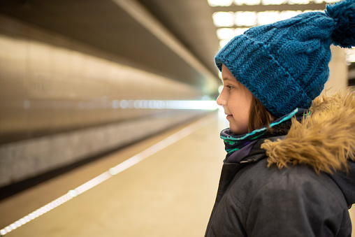 a smiling boy in a winter jacket and hat is waiting for an approaching train on a subway platform