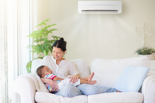Mother and baby under cool air conditioner. Comfortable temperature at family home. Cooling and heating device. Asian mom and kid on couch under cold breeze. Air conditioning on hot summer day.