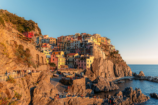 a breathtaking sunset view of the charming village of Manarola in Italy's 5 Terre region. The warm golden hues of the sun perfectly complement the colorful buildings, creating a stunning contrast against the deep blue waters of the Mediterranean Sea.