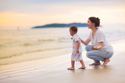Mother and baby on tropical beach at sunset. Asian woman with infant boy walking and playing at sea. Young mom and little boy walk on ocean coast. Travel with kids. Vacation with young child.