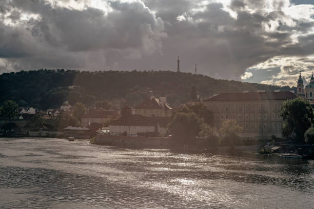 A panoramic view from the historic Charles Bridge, overlooking the Vltava River and the iconic Prague riverfront on a cloudy day. In the distance, the Legion Bridge (Most legií) spans the river, connecting Prague’s key districts. The riverbank is lined with a mix of architectural styles, from neo-Renaissance and Baroque buildings to Art Nouveau and early 20th-century constructions, reflecting Prague’s rich history and diverse design influences. The overcast sky adds a moody atmosphere to the timeless beauty of the cityscape.