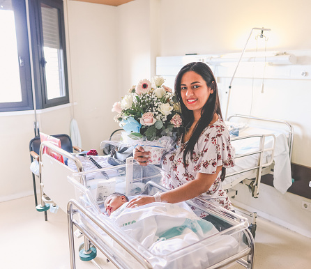 latina woman next to her newborn baby in a hospital, smiling and holding a bouquet of flowers.