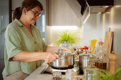 Portrait of mature smiling woman holding a saucepan on glass-ceramic stove top while cooking Italian spaghetti